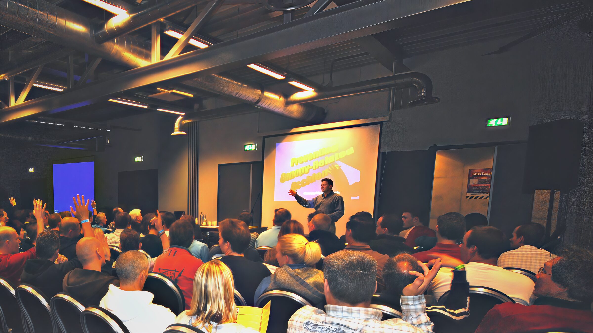 A packed hall seen from the back, hands raised, Brian Germain speaking at the front